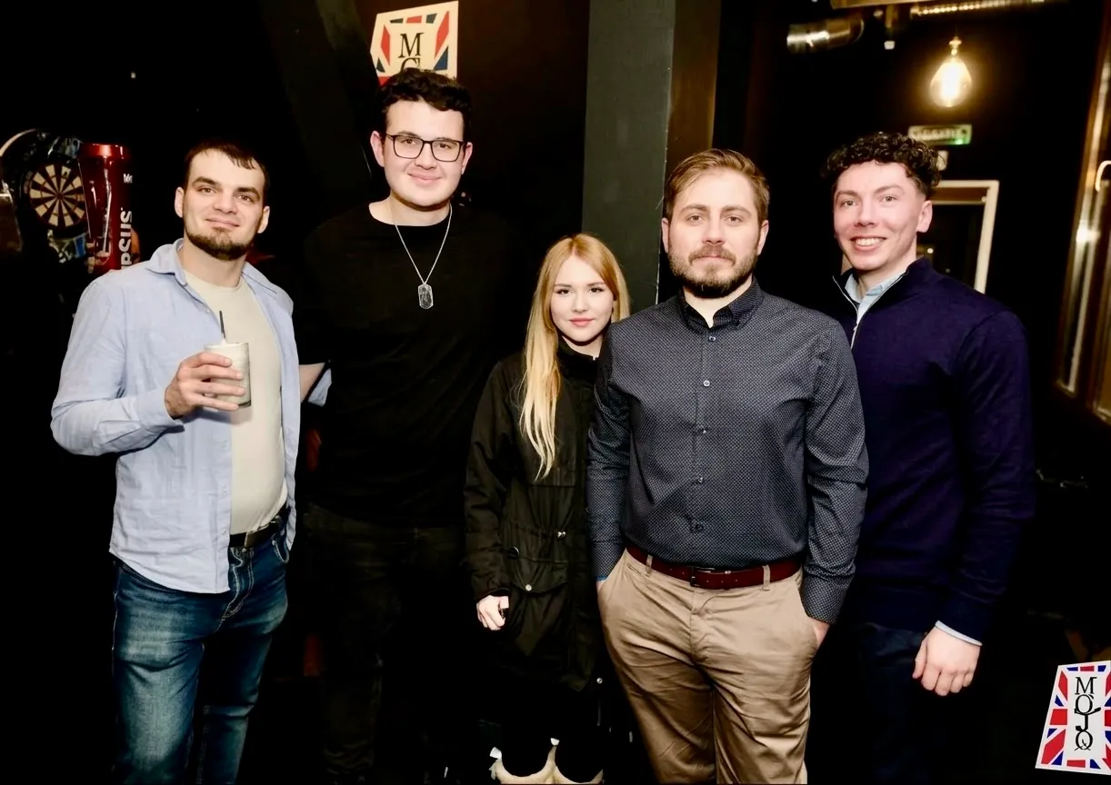 Cinci persoane în picioare, zâmbind, la un eveniment Gatherings într-un local cu lumină caldă.